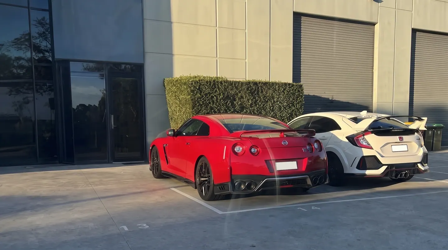 A red Nissan GT-R and a white Honda Civic Type R parked side by side outside an industrial unit in the afternoon sun.