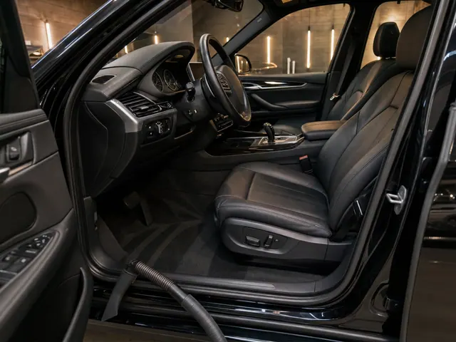 The cabin of a BMW X5 from the open driver's door, the seat and carpet freshly cleaned, a vacuum hose resting on the sill.