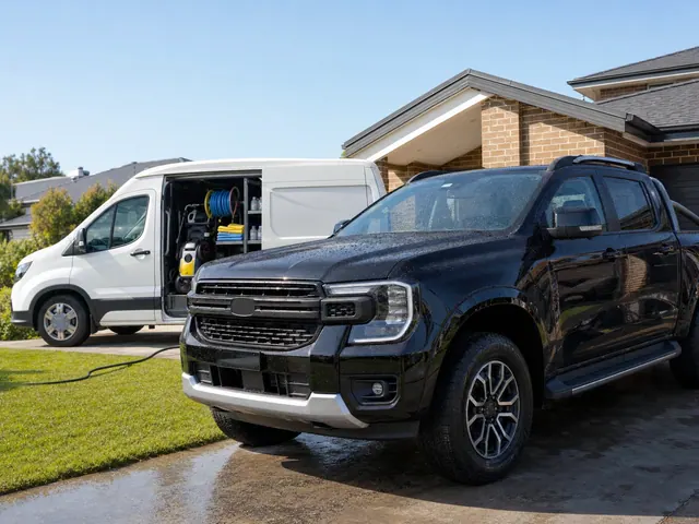 A plain white van with its side door open on a pressure washer and a hose reel, beside a freshly washed black Ford Ranger in a suburban driveway.